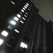 photo of a tall apartment building on a gray night. some of the square windows are illuminated. the building looms above the viewer